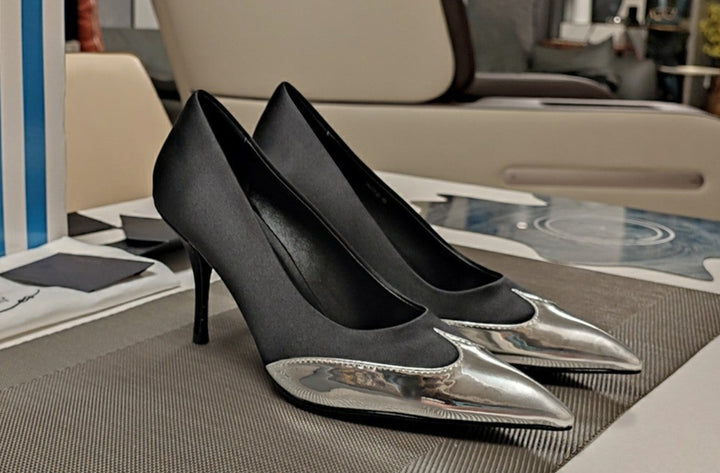 Black high-heeled shoes with silver tips on a table next to a Prada bag.